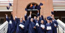 Sixteen students, all wearing blue robes, throw their mortar-board hats in the air as they stand before the brick entrance to Old Main.
