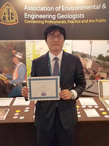 A man standing before a table and a banner reading "Association of Environmental and Engineering Geologists; Connecting Professionals, Practice and the Public" while holding up a certificate. 