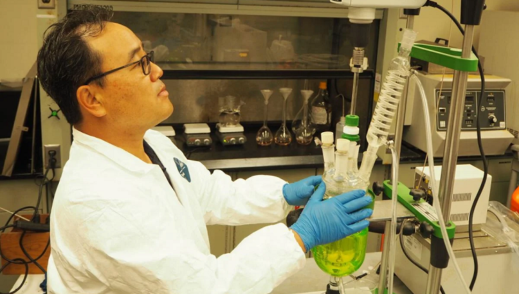 Jaeheon Lee wears a lab coat and latex gloves while holding a green beaker containing lab samples.