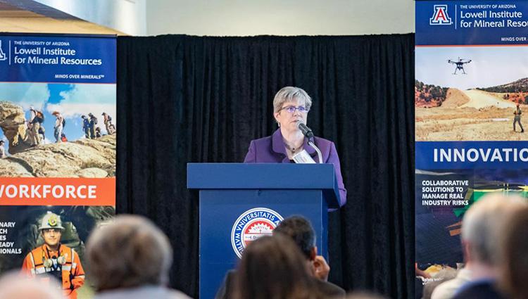 Mary Poulton stands behind a blue podium with the red-and-blue University of Arizona seal on it. She is flanked on either side by large posters touting the Lowell Institute's strengths in workforce development and innovation. 